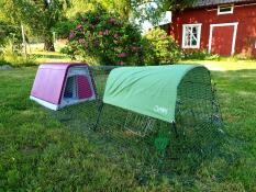 A pink coop with run attached, covered by a shade, with a beautiful red house in the background