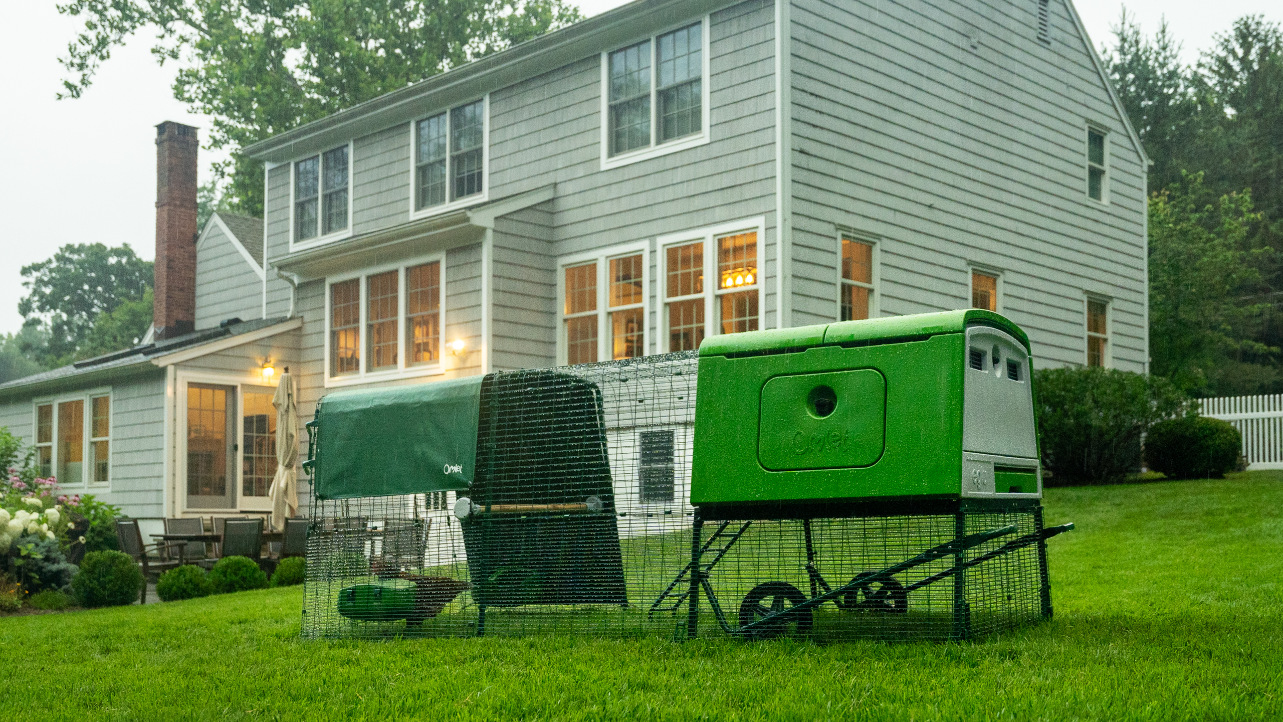 Eglu Cube chicken coop and run in a garden during rainfall, in front of a large house